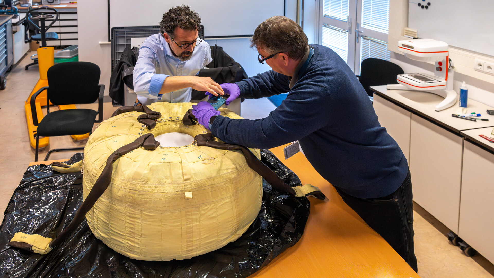 Two people wearing glasses work above what looks like a large tire with fabric on the outside on a table. 