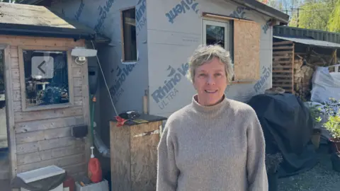 Jane Coyle, a woman in her 60s, is standing in front of a shed made of chipboard