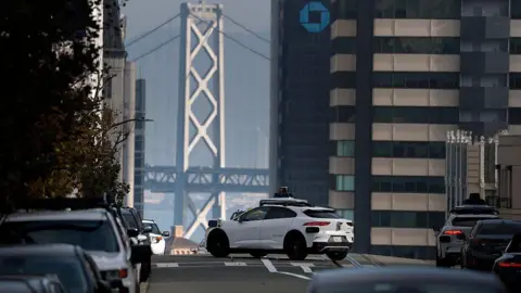 Getty Images A white autonomous robotaxi driving on a city street with the Golden Gate Bridge and San Francisco Bay in the background. There are cars parked on the side of the street. 