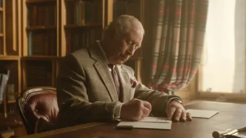 King Charles III writes a letter from a desk in front of a window in Buckingham Palace. He is wearing a blazer and a purple tie. 