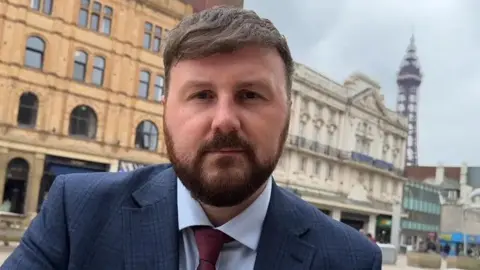 Head and shoulders shot of MP Chris Webb in Blackpool. He has short dark hair and a beard and is wearing a dark blue suit, pale blue shirt and burgundy tie. He is standing outside with hotels and Blackpool Tower in the background