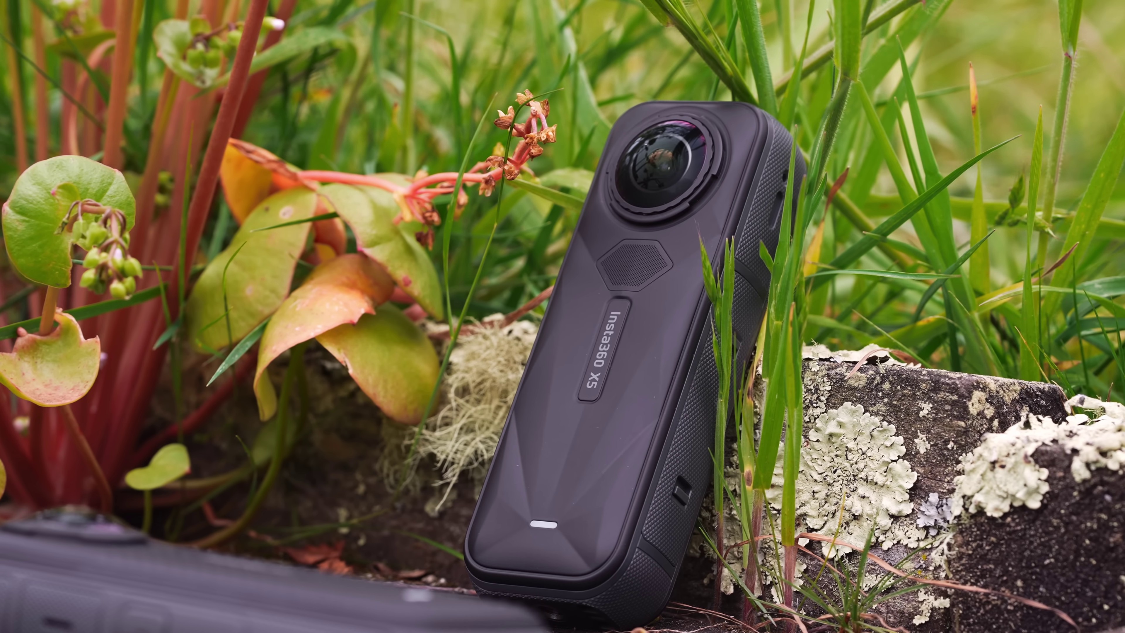 The Insta360 X5 360-degree black stick camera sitting in a bed of flowers and grass.